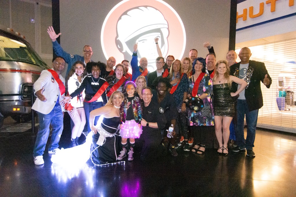 A diverse group of aerospace supporters wearing red sashes cheer in front of a projected Yuri's Night logo (a smiling astronaut head)