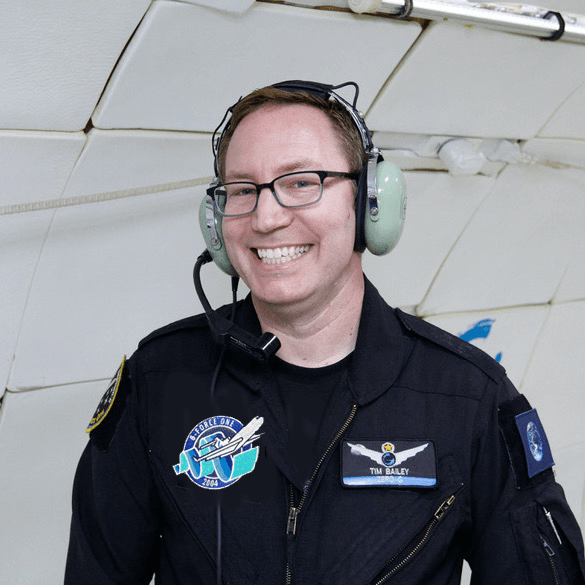 Tim Bailey, smiling, stands inside the padded fuselage of a parabolic aircraft while wearing a black flight suit and green aviation headset.